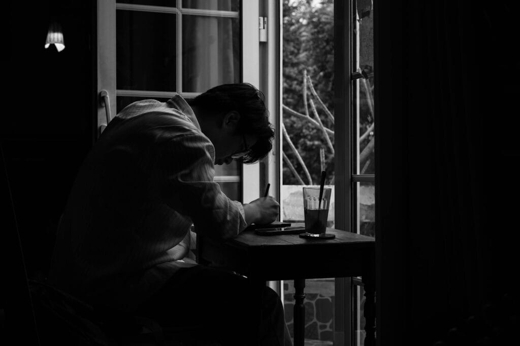 A person writes at a table near a window.