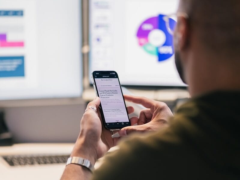 Man using smartphone in front of computer monitors.